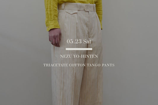 根津洋品店（NEZU YO-HINTEN）のTANGO PANTS（タンゴパンツ）を着用した男性（身長184cm）の全身カット。2026年パントン・カラー「クラウドダンサー」を捉えたライトイエロー（クリーム）の色相。トリアセテート高密度ジャガードが生む立体的な無地ストライプのテクスチャーと、前後タックが描く極太の円柱シルエット。中央に白いブランドロゴテキスト。