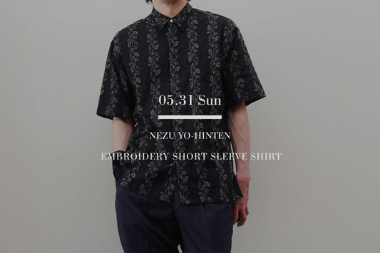 根津洋品店のEMBROIDERY SHORT SLEEVE SHIRTを着用した男性のスタイリング画像。黒のコットン生地に同系色で植物調の総刺繍が施されたフロント比翼仕様の半袖ドレスシャツとネイビーのトラウザーズのコーディネート。中央に白いブランドロゴテキスト。