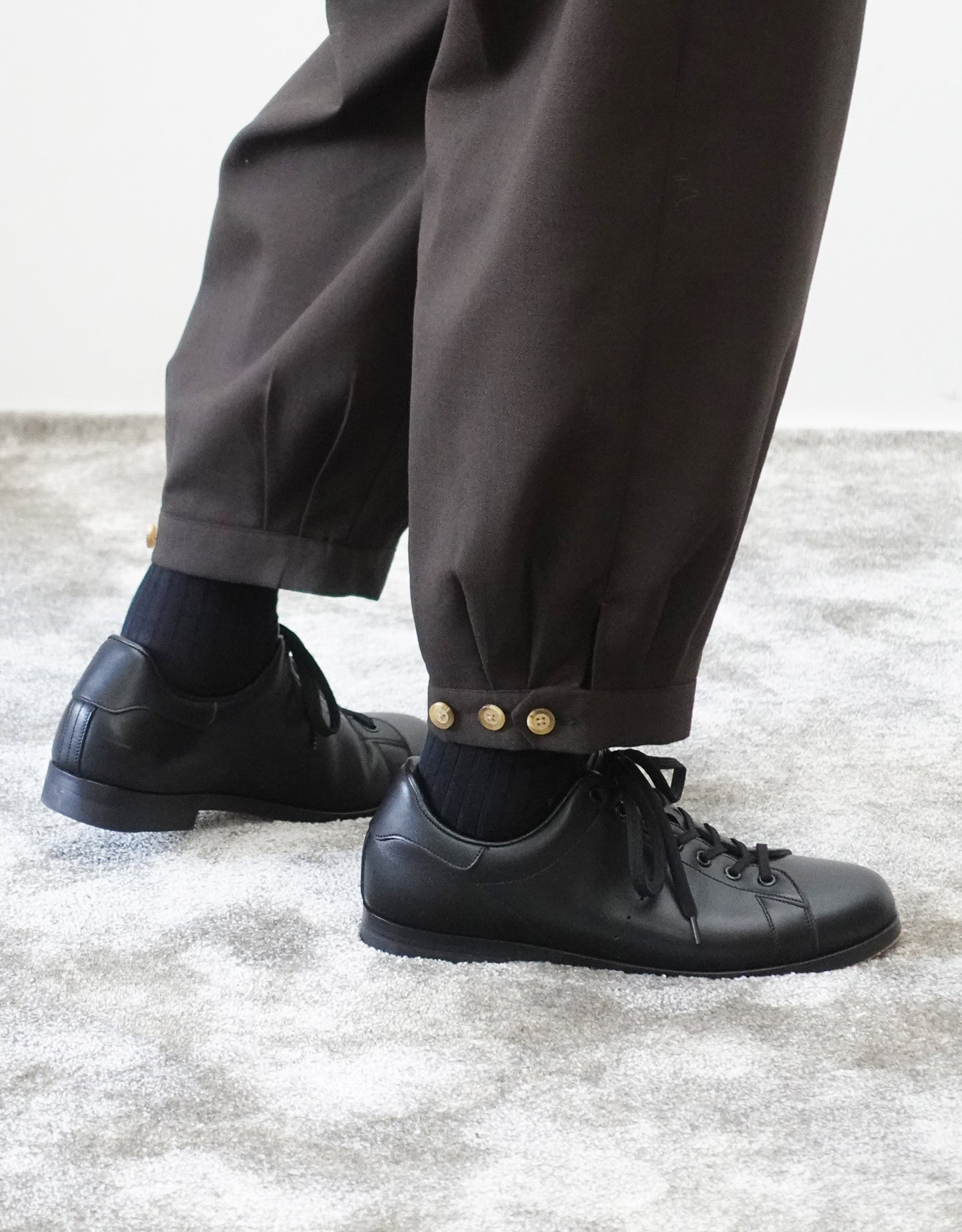 VOUILLON（ヴイヨン）のTriple Tuck Harlem Slacks - DARK BROWN（トリプルタックハーレムスラックス）。腰元の3タックによるボリューム感と、裾にかけて絞られたハーレムシルエットが特徴のブラック・ワイドパンツ。裾のエイトタックとシルエットを変更できるボタンがわかる寄り画像。