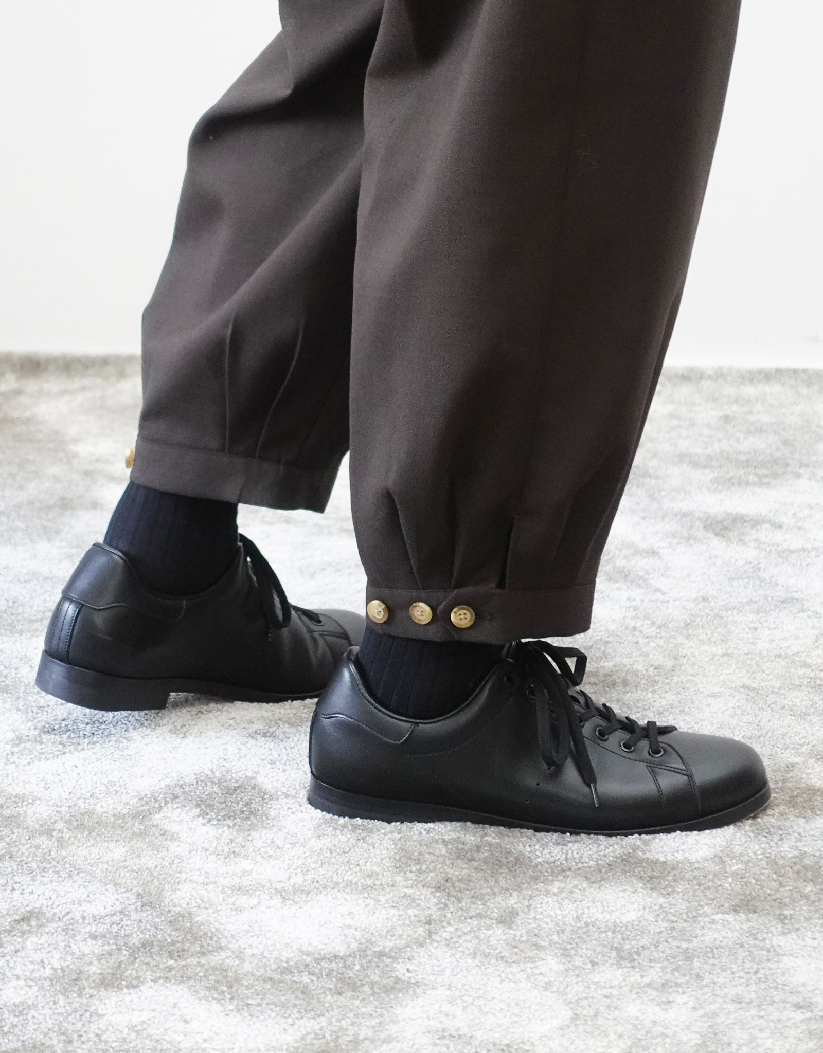 VOUILLON（ヴイヨン）のTriple Tuck Harlem Slacks - DARK BROWN（トリプルタックハーレムスラックス）。腰元の3タックによるボリューム感と、裾にかけて絞られたハーレムシルエットが特徴のブラック・ワイドパンツ。裾のエイトタックとシルエットを変更できるボタンがわかる寄り画像。