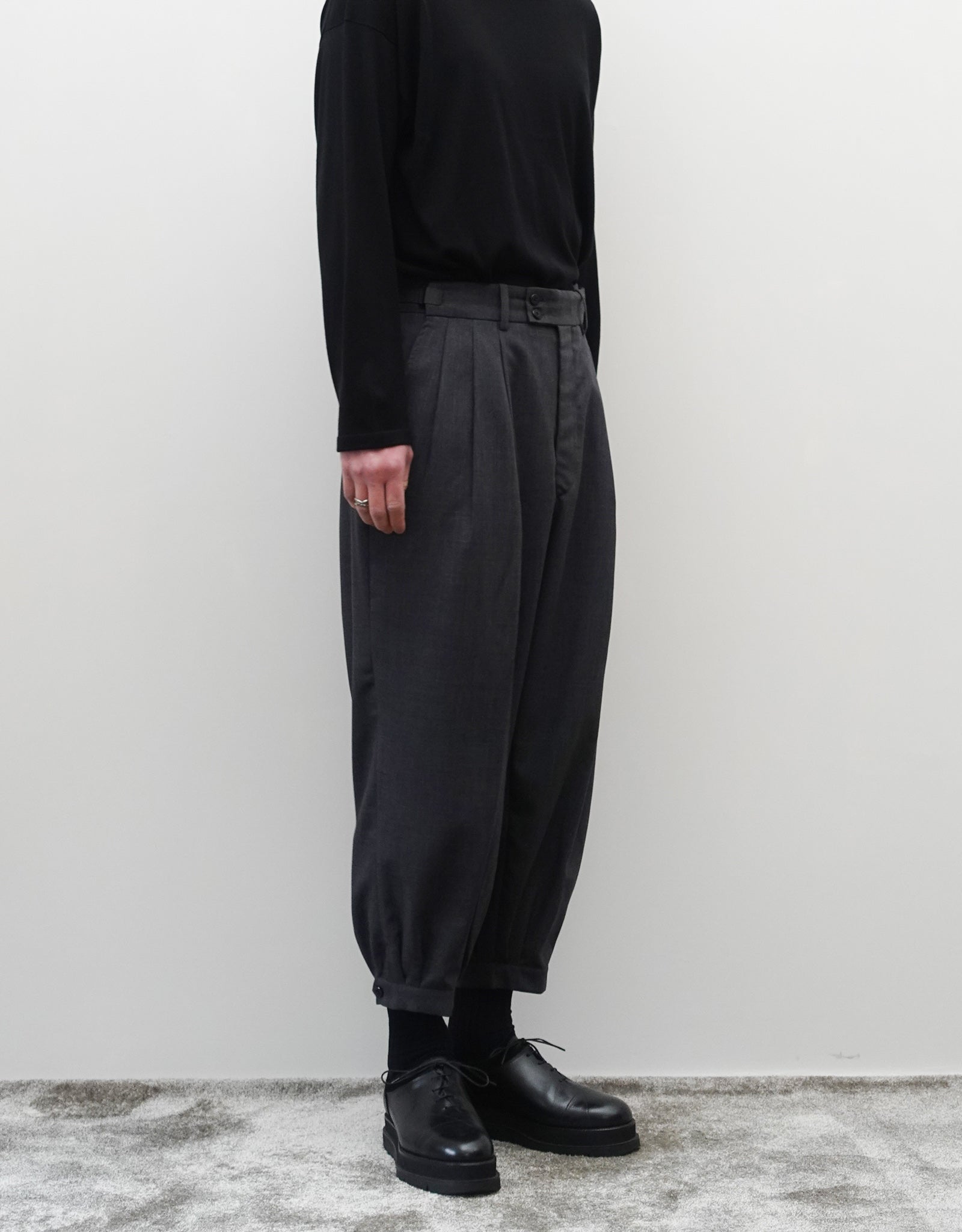 VOUILLON（ヴイヨン）のTriple Tuck Harlem Slacks - DARK GRAY（トリプルタックハーレムスラックス）。腰元の3タックによるボリューム感と、裾にかけて絞られたハーレムシルエットが特徴のダークグレー・ワイドパンツ。左横向きの着用画像。