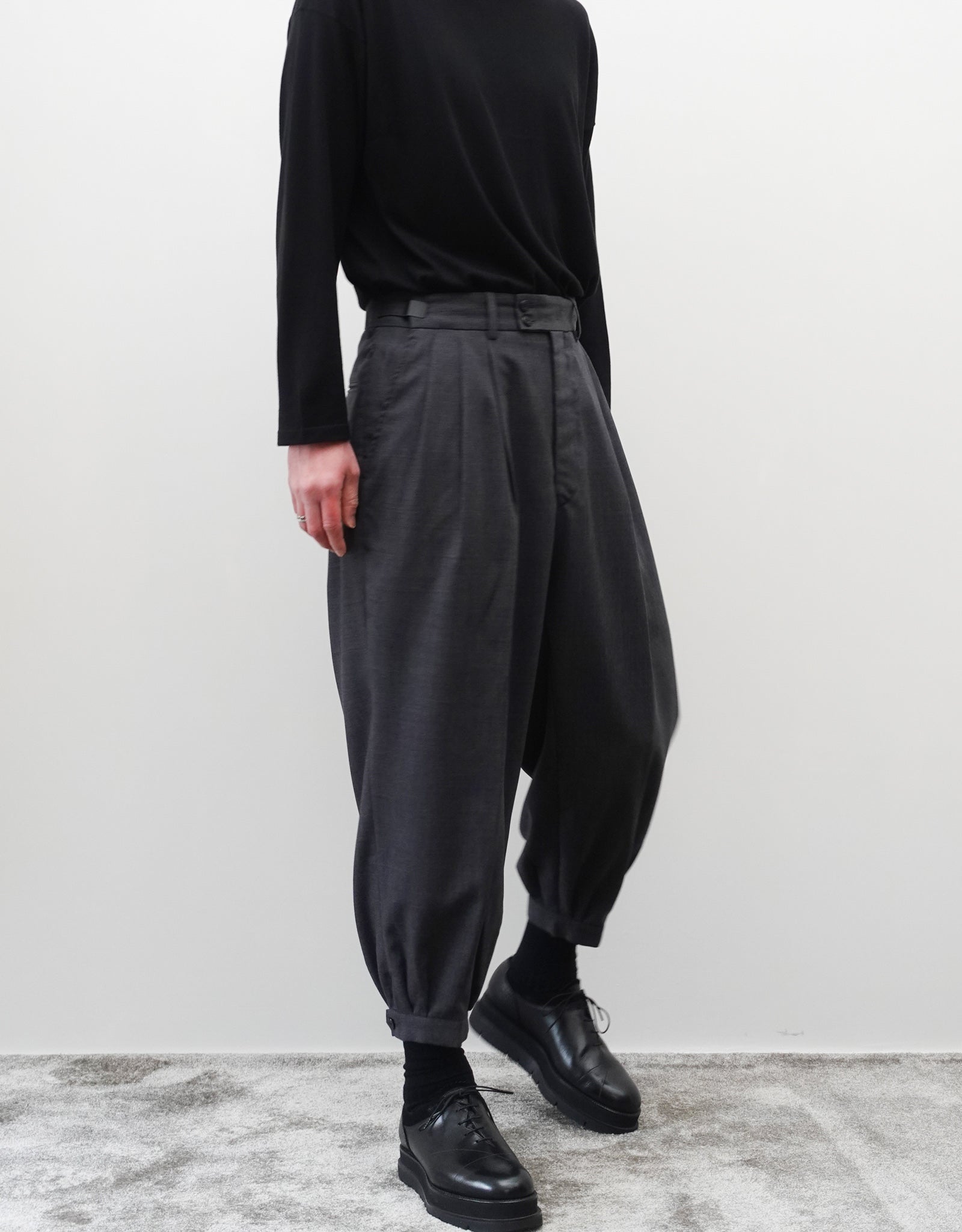 VOUILLON（ヴイヨン）のTriple Tuck Harlem Slacks - DARK GRAY（トリプルタックハーレムスラックス）。腰元の3タックによるボリューム感と、裾にかけて絞られたハーレムシルエットが特徴のダークグレー・ワイドパンツ。左斜め前を向いた首から下の着用画像でシルエットがしっかりわかる。