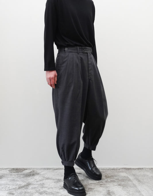 VOUILLON（ヴイヨン）のTriple Tuck Harlem Slacks - DARK GRAY（トリプルタックハーレムスラックス）。腰元の3タックによるボリューム感と、裾にかけて絞られたハーレムシルエットが特徴のダークグレー・ワイドパンツ。左斜め前を向いた首から下の着用画像でシルエットがしっかりわかる。