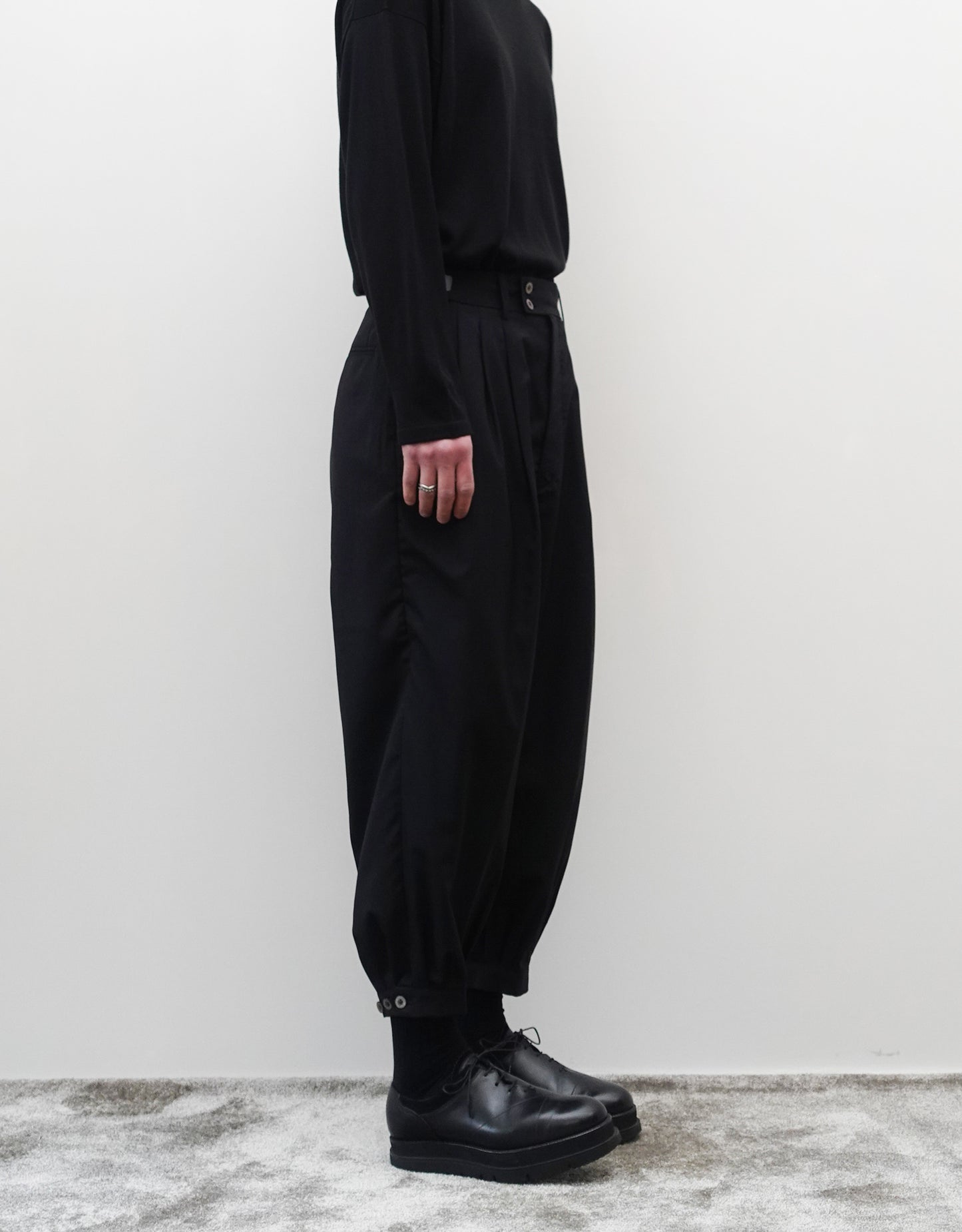 VOUILLON（ヴイヨン）のTriple Tuck Harlem Slacks - Black（トリプルタックハーレムスラックス）。腰元の3タックによるボリューム感と、裾にかけて絞られたハーレムシルエットが特徴のブラック・コクーンシルエットのパンツ。シルエットがわかる左向きの着用画像。