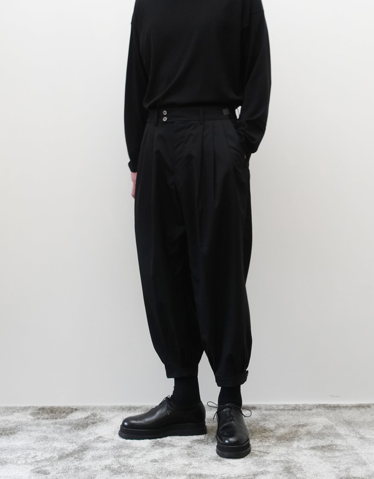 VOUILLON（ヴイヨン）のTriple Tuck Harlem Slacks - Black（トリプルタックハーレムスラックス）。腰元の3タックによるボリューム感と、裾にかけて絞られたハーレムシルエットが特徴のブラック・コクーンシルエットのパンツ。シルエットがわかる正面の着用画像（左手ポケットに手を入れている）。