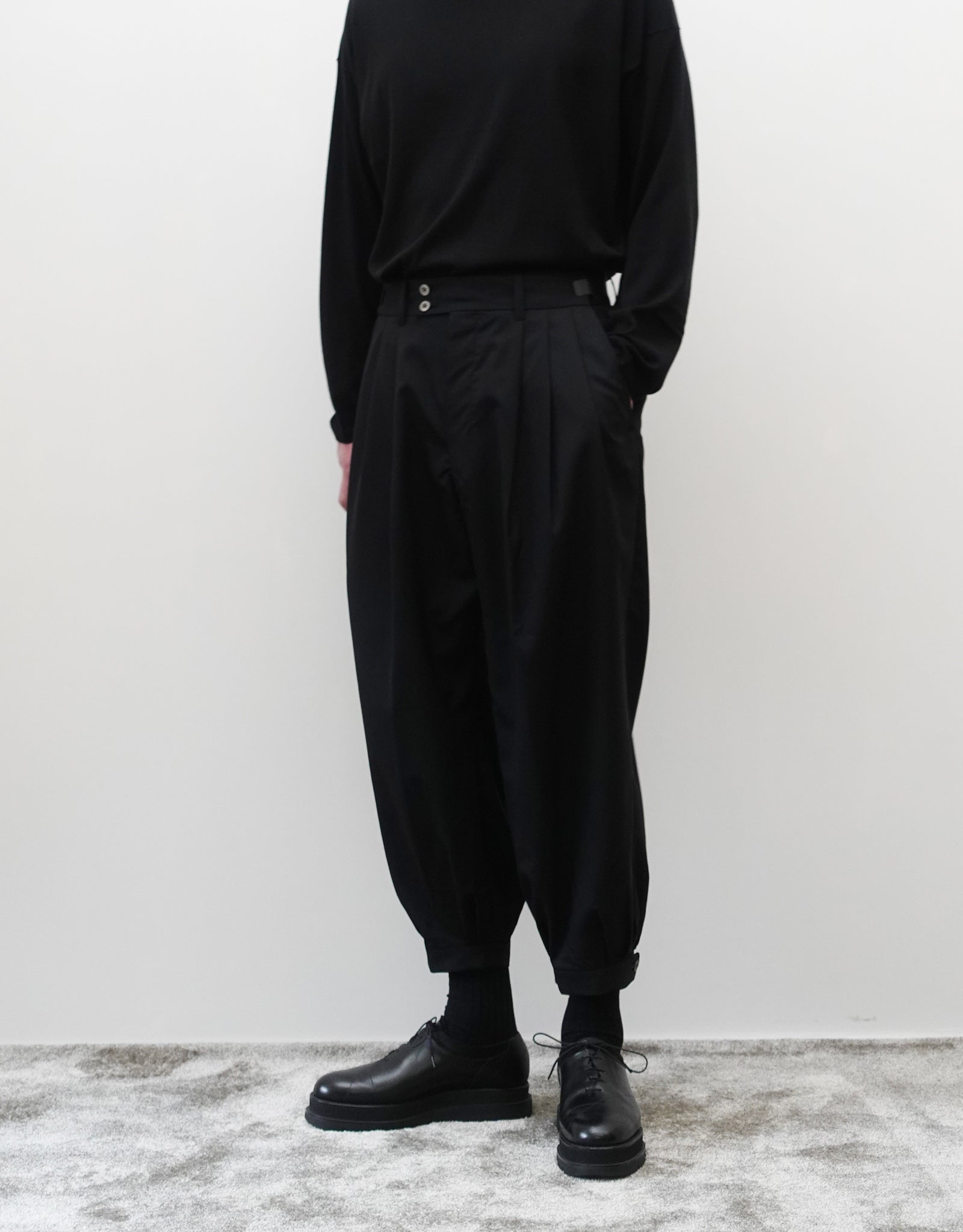 VOUILLON（ヴイヨン）のTriple Tuck Harlem Slacks - Black（トリプルタックハーレムスラックス）。腰元の3タックによるボリューム感と、裾にかけて絞られたハーレムシルエットが特徴のブラック・コクーンシルエットのパンツ。シルエットがわかる正面の着用画像（左手ポケットに手を入れている）。