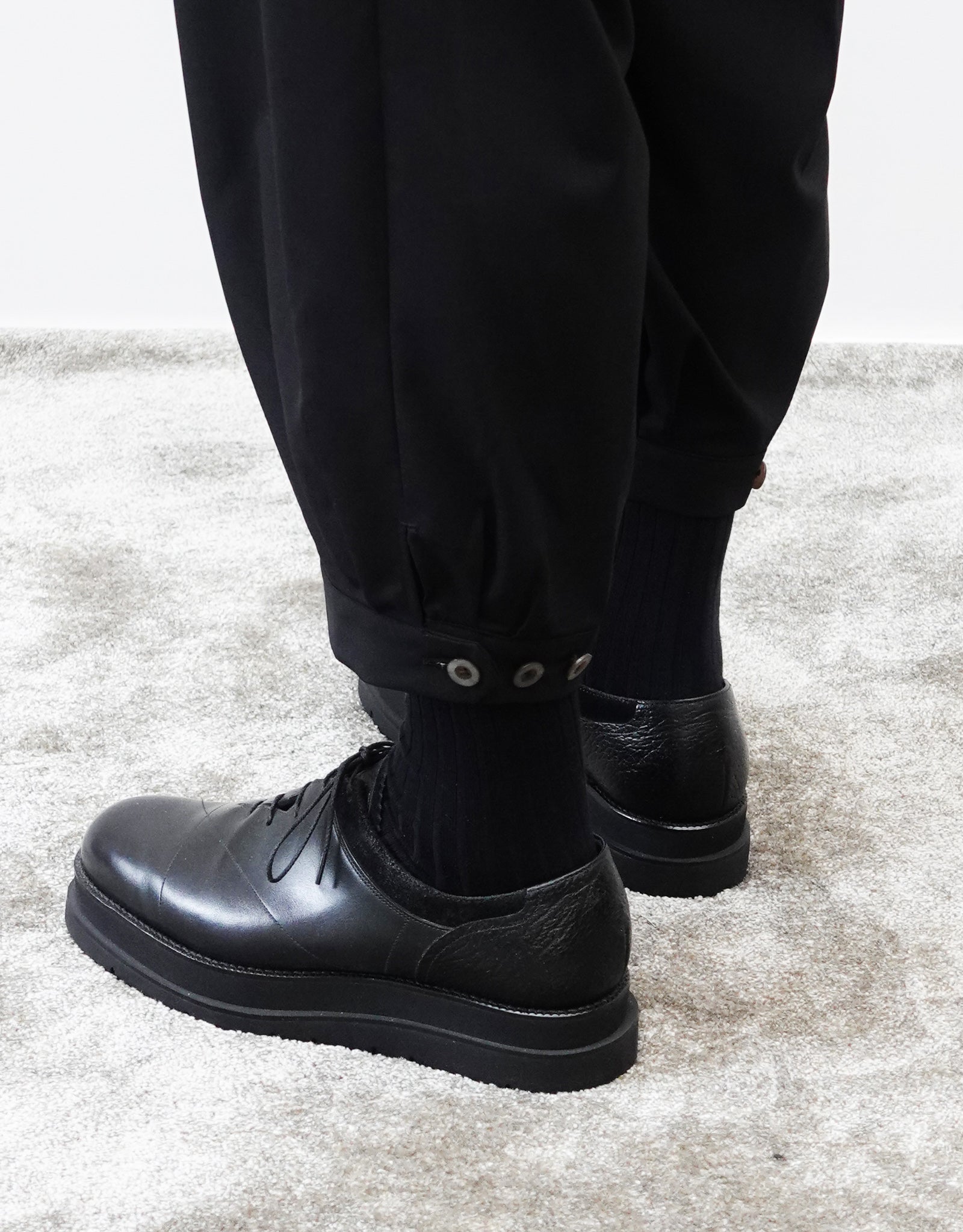 VOUILLON（ヴイヨン）のTriple Tuck Harlem Slacks - Black（トリプルタックハーレムスラックス）。腰元の3タックによるボリューム感と、裾にかけて絞られたハーレムシルエットが特徴のブラック・コクーンシルエットのパンツ。裾のエイトタックをアップにした背面から撮影。