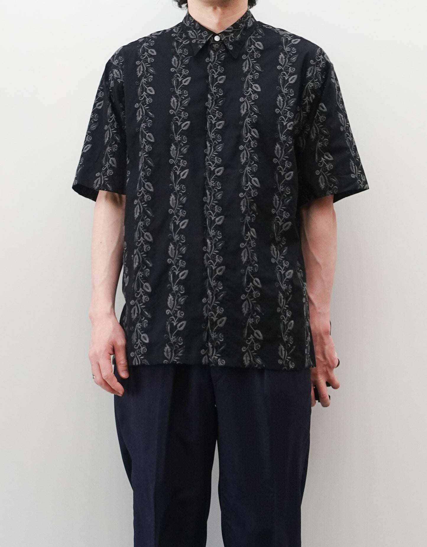 NEZU YO-HINTEN（根津洋品店）のEmbroidery Short Sleeve Shirt - Black（エンブロイダリー半袖シャツ）。前身頃に施された植物モチーフの垂直な刺繍が特徴。ブラックの生地にグレーの刺繍が映える、モードなリラックスフィットのスタイリング。モデル正面着用画像。