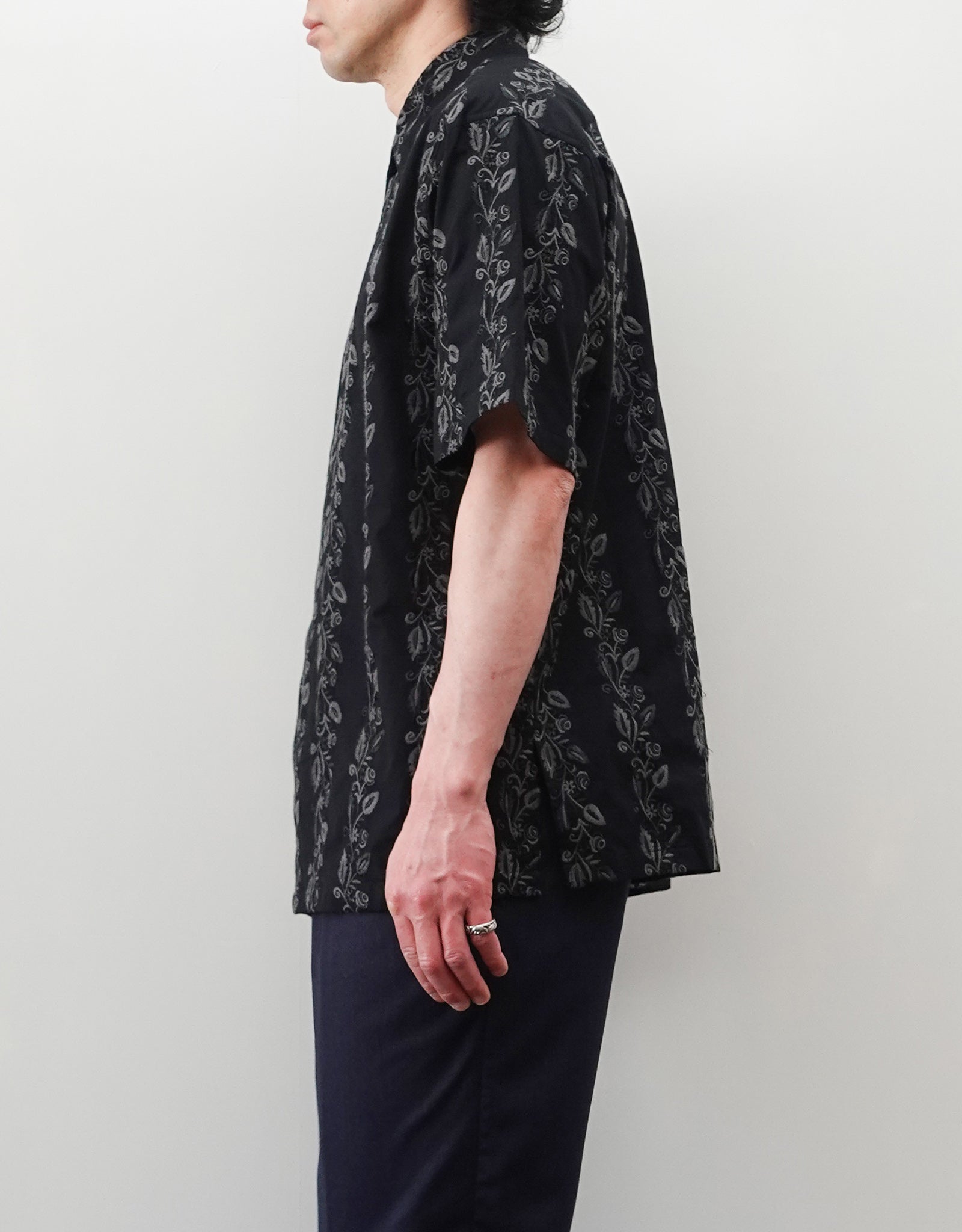 NEZU YO-HINTEN（根津洋品店）のEmbroidery Short Sleeve Shirt - Black（エンブロイダリー半袖シャツ）。前身頃に施された植物モチーフの垂直な刺繍が特徴。ブラックの生地にグレーの刺繍が映える、モードなリラックスフィットのスタイリング。モデル右向き着用画像。