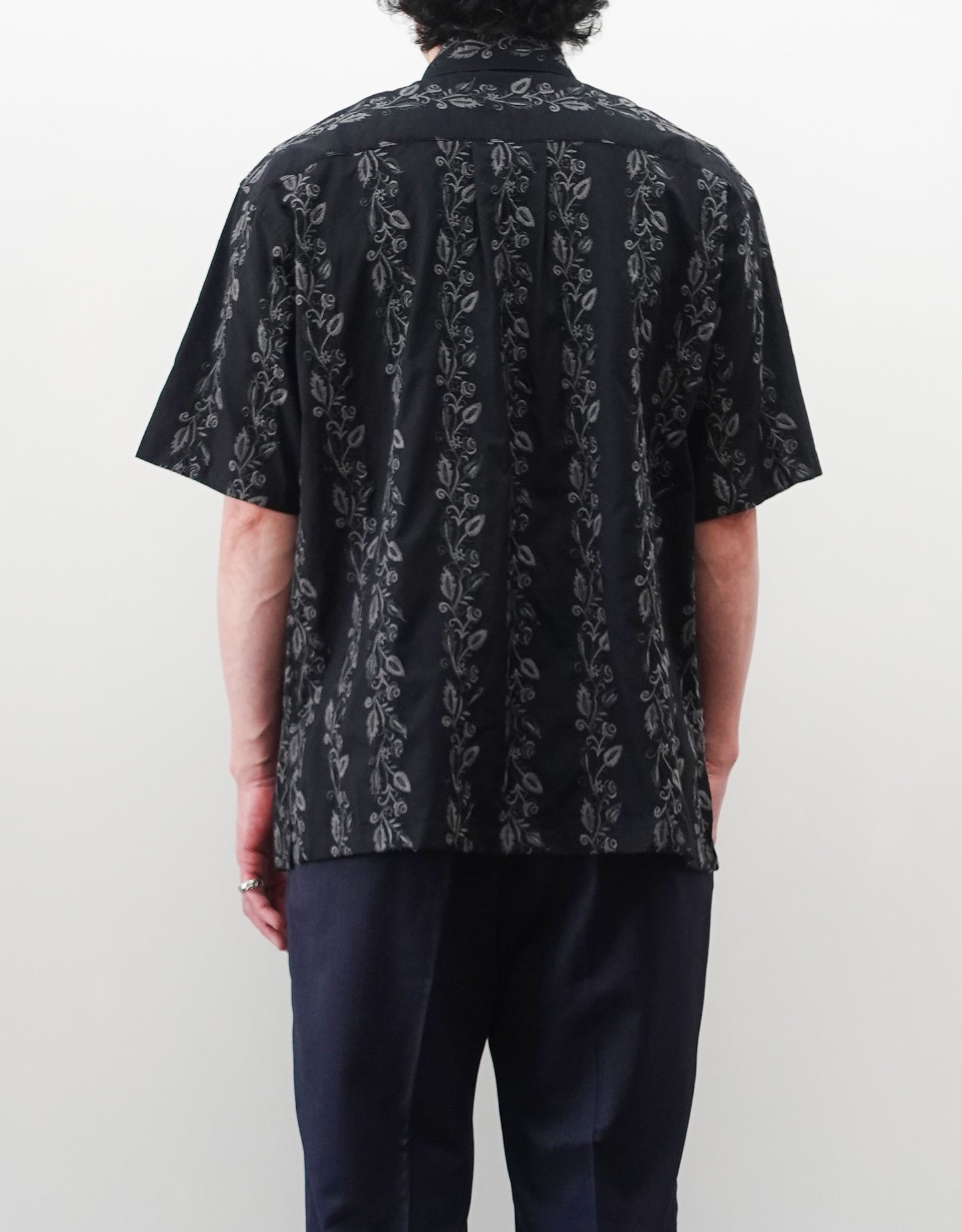 NEZU YO-HINTEN（根津洋品店）のEmbroidery Short Sleeve Shirt - Black（エンブロイダリー半袖シャツ）。前身頃に施された植物モチーフの垂直な刺繍が特徴。ブラックの生地にグレーの刺繍が映える、モードなリラックスフィットのスタイリング。モデル背面着用画像。