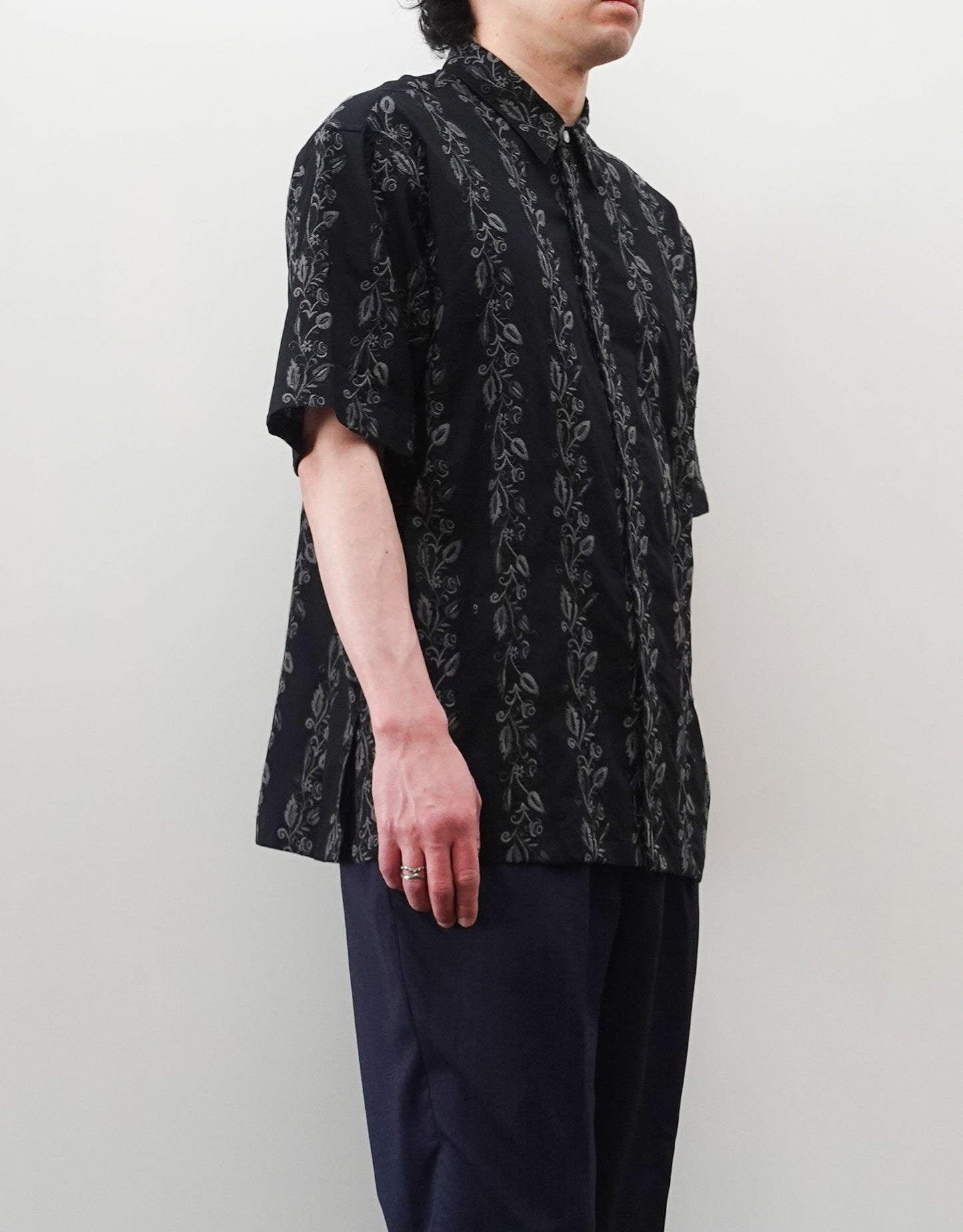 NEZU YO-HINTEN（根津洋品店）のEmbroidery Short Sleeve Shirt - Black（エンブロイダリー半袖シャツ）。前身頃に施された植物モチーフの垂直な刺繍が特徴。ブラックの生地にグレーの刺繍が映える、モードなリラックスフィットのスタイリング。モデル左向き着用画像。