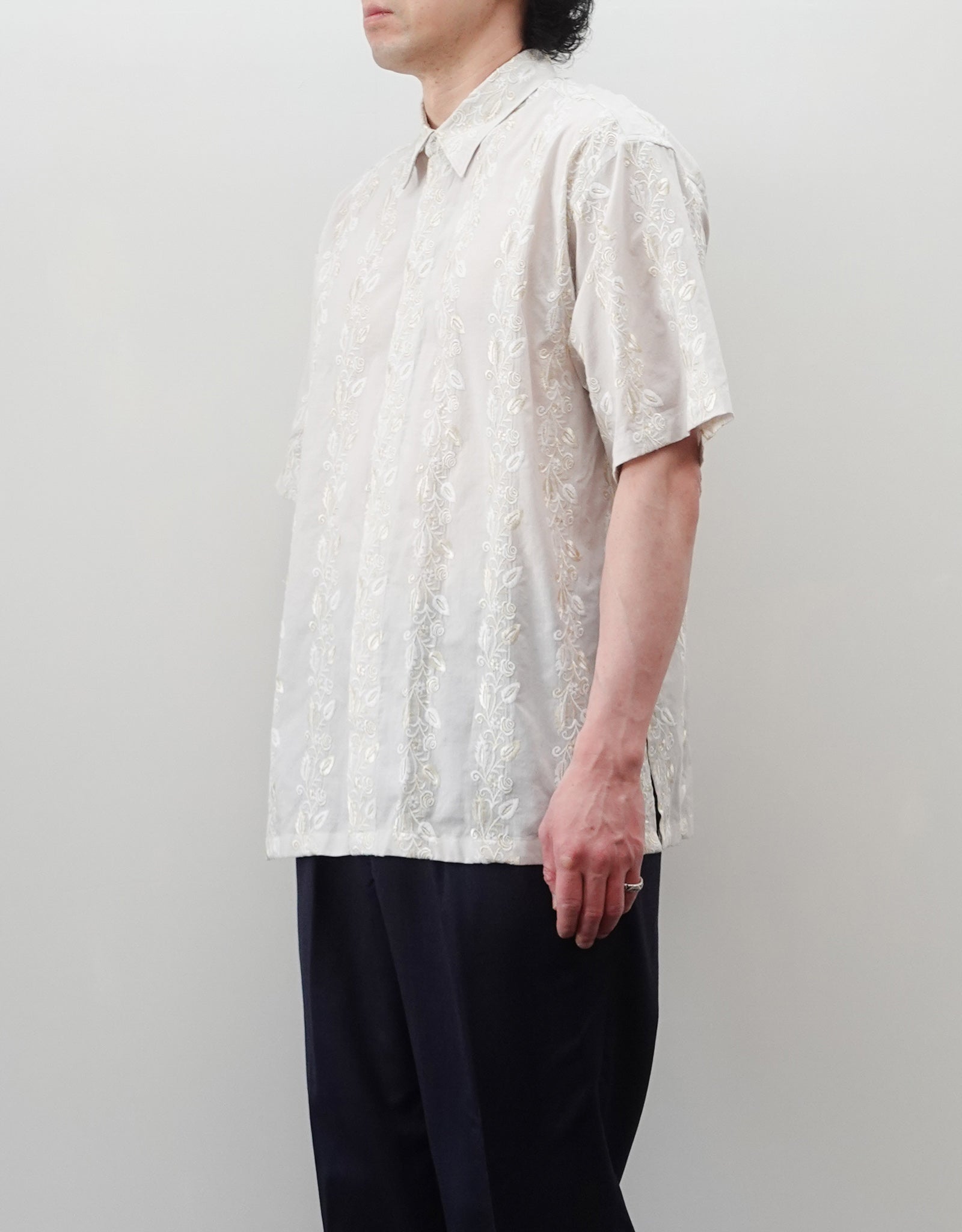 NEZU YO-HINTEN（根津洋品店）のEmbroidery Short Sleeve Shirt - Off White（エンブロイダリー半袖シャツ）。前身頃に施された植物モチーフの垂直な刺繍が特徴。ブラックの生地にグレーの刺繍が映える、モードなリラックスフィットのスタイリング。モデル右向き着用画像。