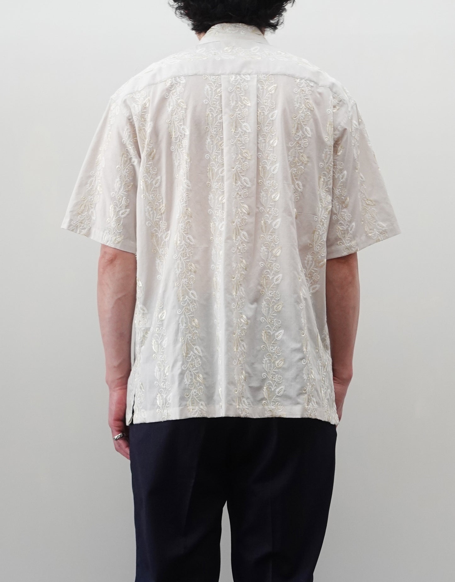 NEZU YO-HINTEN（根津洋品店）のEmbroidery Short Sleeve Shirt - Off White（エンブロイダリー半袖シャツ）。前身頃に施された植物モチーフの垂直な刺繍が特徴。ブラックの生地にグレーの刺繍が映える、モードなリラックスフィットのスタイリング。モデル背面着用画像。