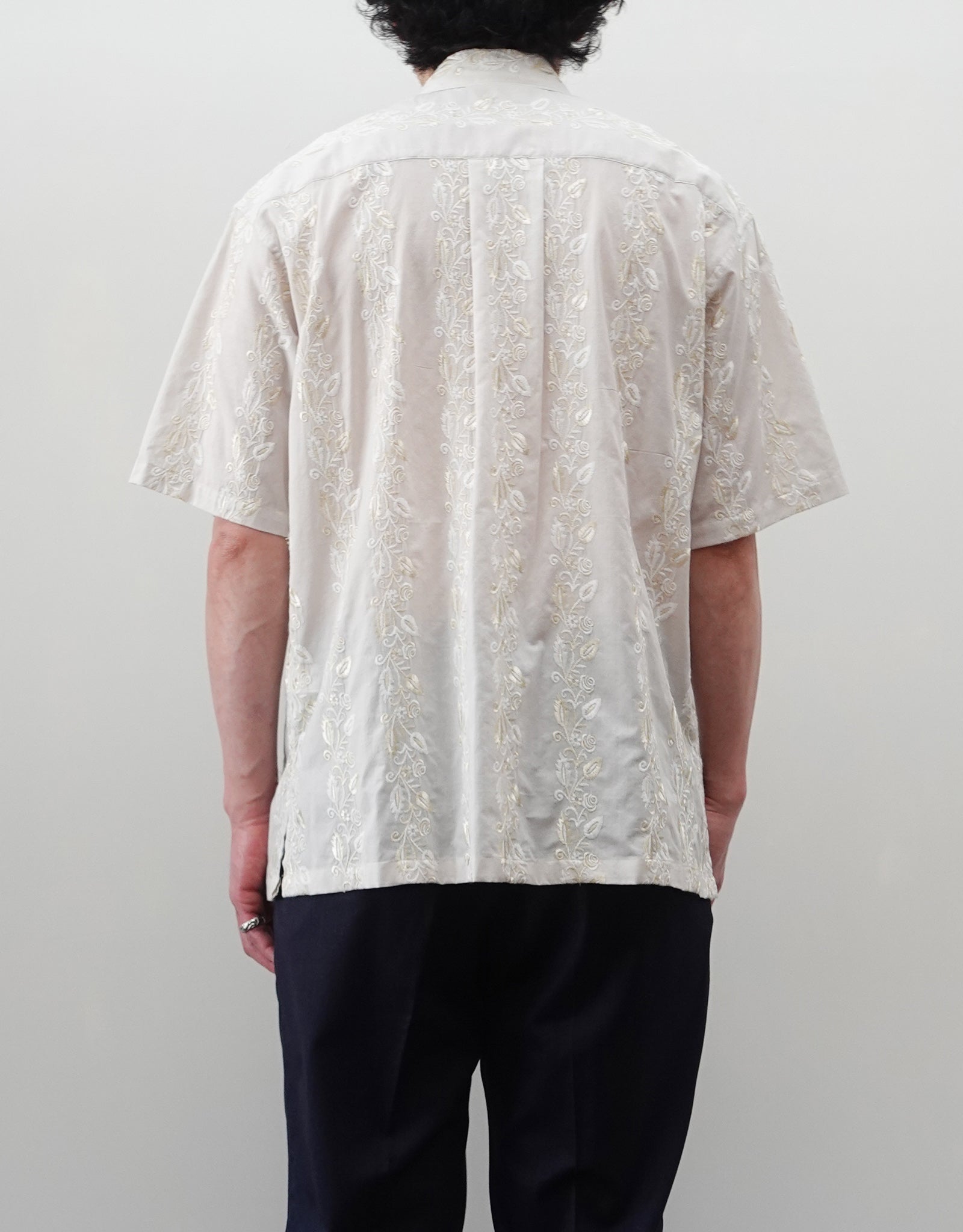 NEZU YO-HINTEN（根津洋品店）のEmbroidery Short Sleeve Shirt - Off White（エンブロイダリー半袖シャツ）。前身頃に施された植物モチーフの垂直な刺繍が特徴。ブラックの生地にグレーの刺繍が映える、モードなリラックスフィットのスタイリング。モデル背面着用画像。