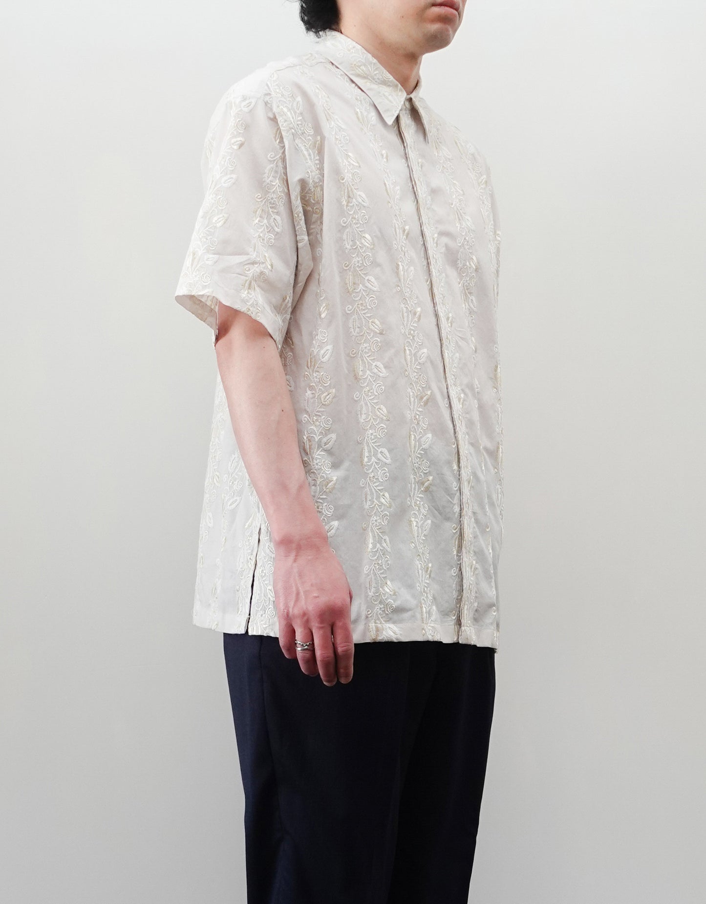 NEZU YO-HINTEN（根津洋品店）のEmbroidery Short Sleeve Shirt - Off White（エンブロイダリー半袖シャツ）。前身頃に施された植物モチーフの垂直な刺繍が特徴。ブラックの生地にグレーの刺繍が映える、モードなリラックスフィットのスタイリング。モデル左向き着用画像。