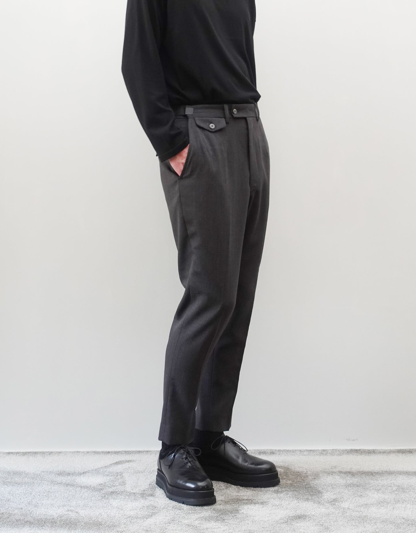 VOUILLON（ヴイヨン）のTwo Darts Slim Trousers - Gray Multi Stripe（2ダーツスリムトラウザーズ）。グレーマルチストライプ柄の端正な生地に、ウエスト回りをすっきり見せる2ダーツ仕様のスリムパンツ。美しいスリムテーパードシルエット。モデル着用左斜め前向き画像。