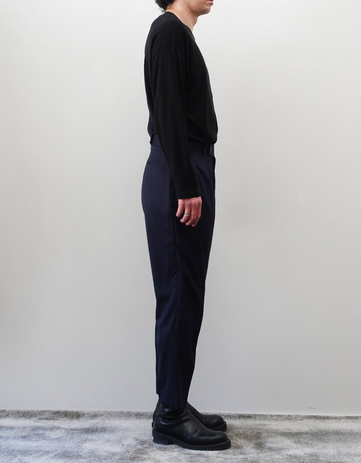 VOUILLON（ヴイヨン）のSuspender Two Tuck Trousers - Navy（サスペンダーツータックトラウザーズ）。ネイビーのツータックワイドパンツに、取り外し可能な同素材のサスペンダーを備えたディテール。モデル着用左向き画像。