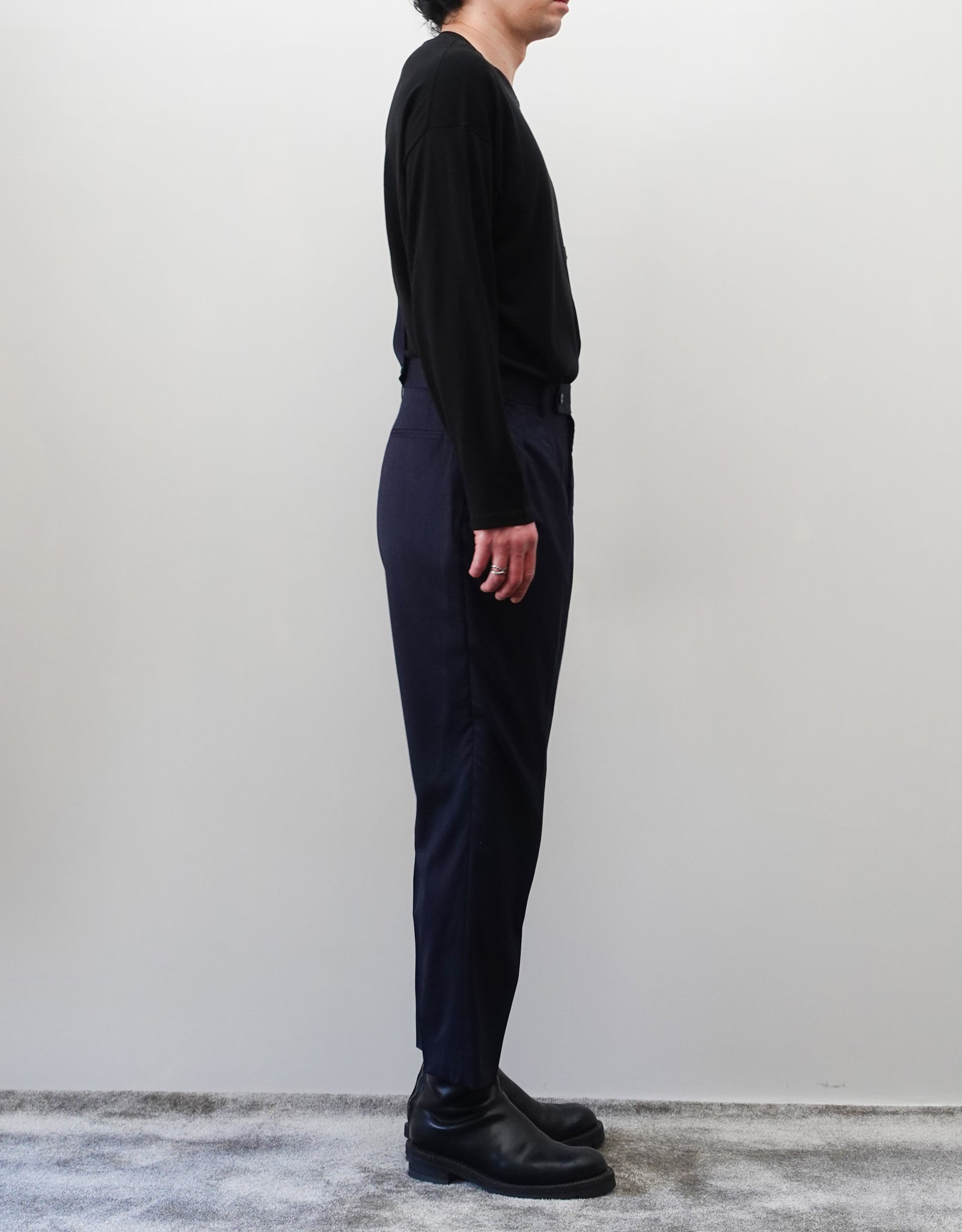VOUILLON（ヴイヨン）のSuspender Two Tuck Trousers - Navy（サスペンダーツータックトラウザーズ）。ネイビーのツータックワイドパンツに、取り外し可能な同素材のサスペンダーを備えたディテール。モデル着用左向き画像。