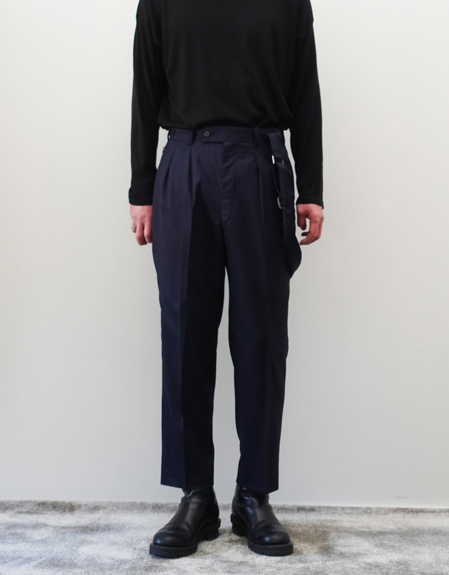 VOUILLON（ヴイヨン）のSuspender Two Tuck Trousers - Navy（サスペンダーツータックトラウザーズ）。ネイビーのツータックワイドパンツに、取り外し可能な同素材のサスペンダーを備えたディテール。モデル着用正面でサスペンダーを肩から外した画像。