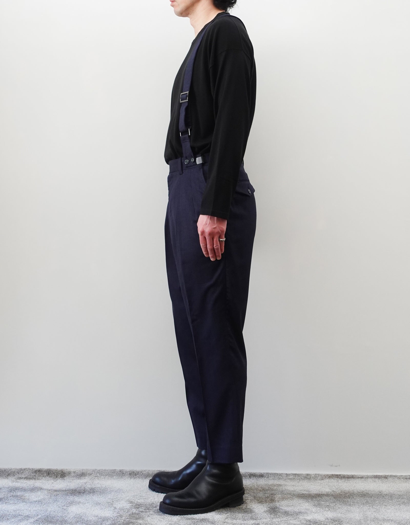VOUILLON（ヴイヨン）のSuspender Two Tuck Trousers - Navy（サスペンダーツータックトラウザーズ）。ネイビーのツータックワイドパンツに、取り外し可能な同素材のサスペンダーを備えたディテール。モデル着用右向き画像。