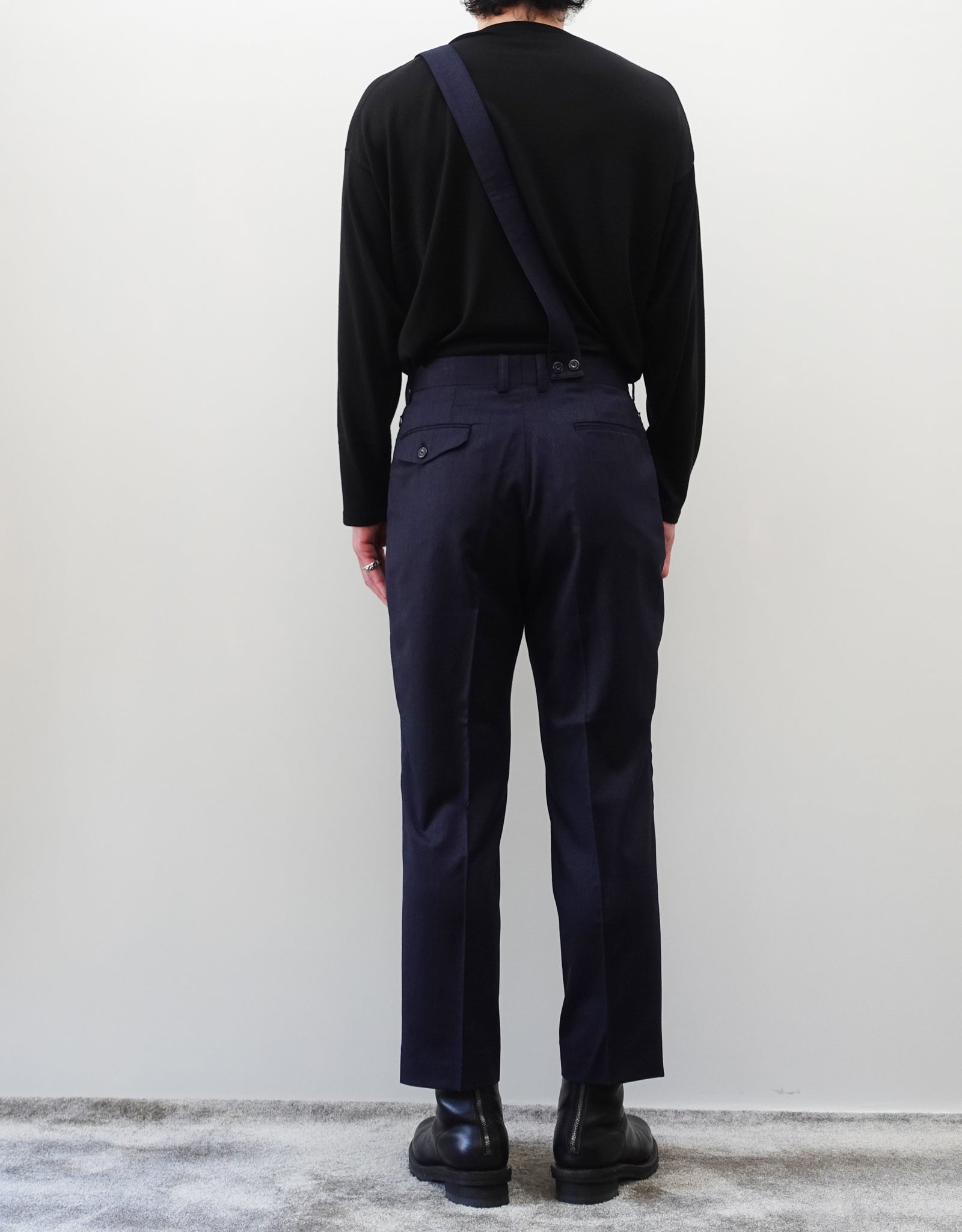 VOUILLON（ヴイヨン）のSuspender Two Tuck Trousers - Navy（サスペンダーツータックトラウザーズ）。ネイビーのツータックワイドパンツに、取り外し可能な同素材のサスペンダーを備えたディテール。モデル着用背面画像。
