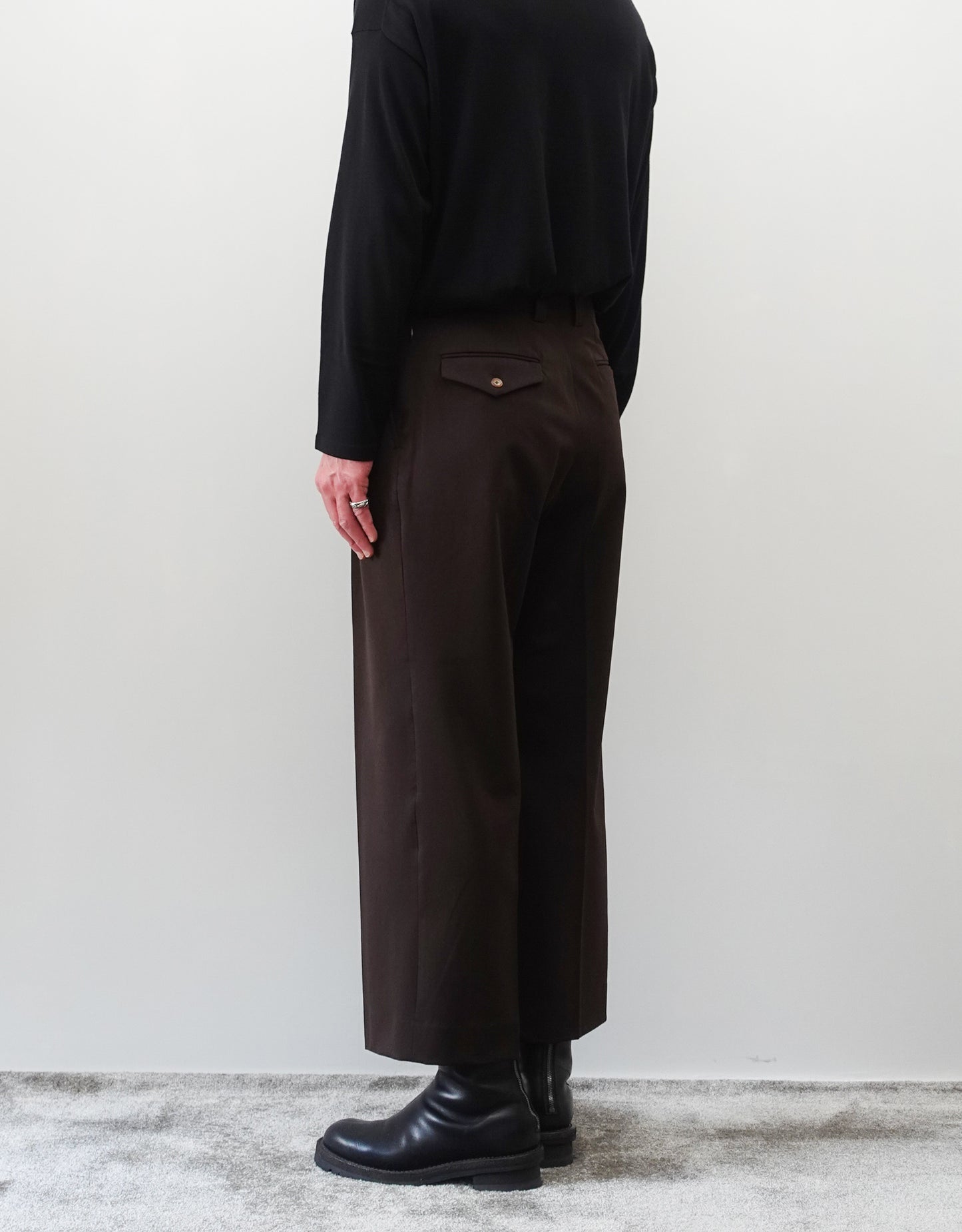 VOUILLON（ヴイヨン）のFour Pleats Wide Trousers - Dark Chocolate（4プリーツワイドトラウザーズ）。ダークチョコレート（深みのあるブラウン）のワイドシルエットと腰元の4プリーツ。モデルによる右斜め後向きの着用画像。