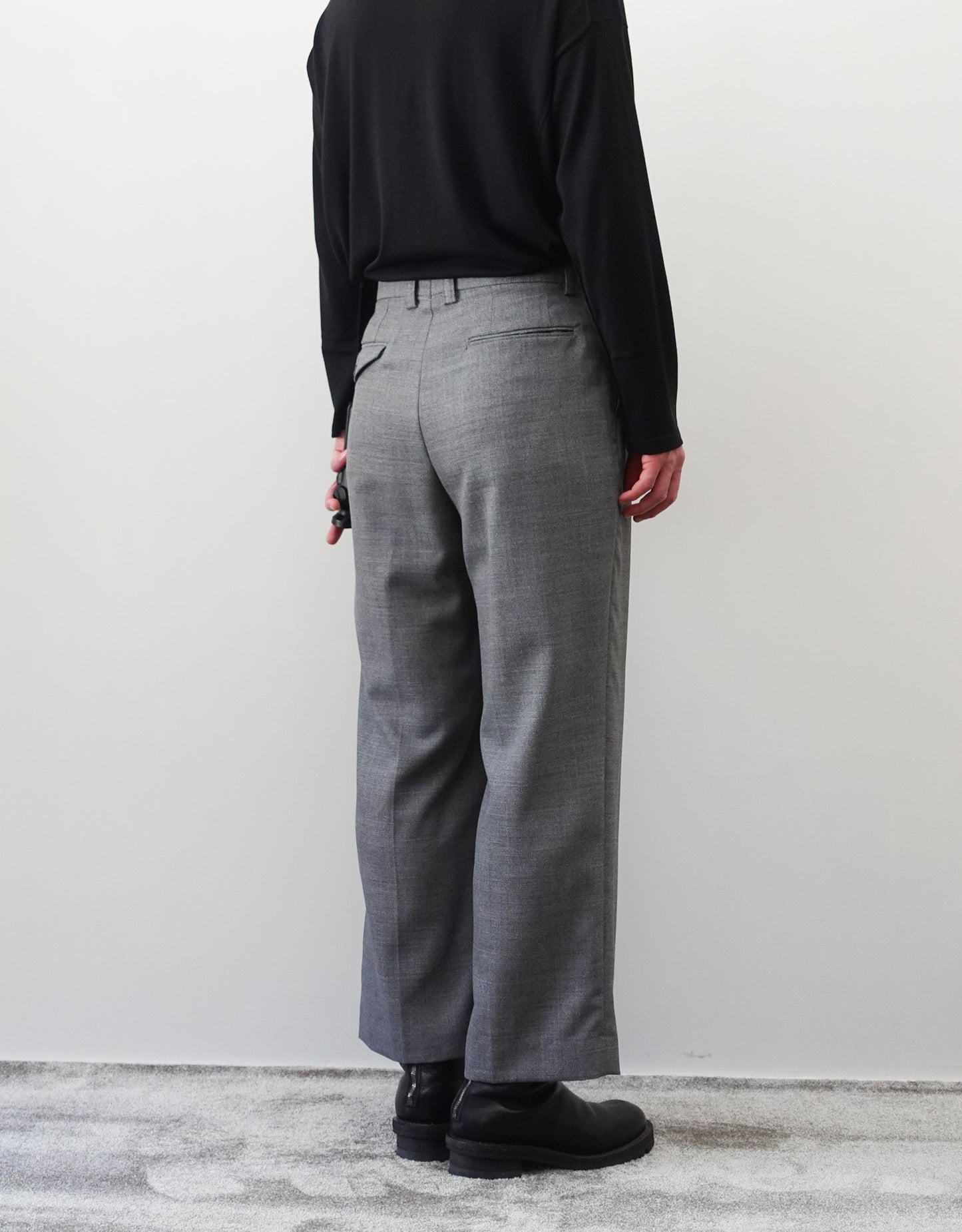 VOUILLON（ヴイヨン）のFour Pleats Wide Trousers - Gray（4プリーツワイドトラウザーズ）。ソリッドなグレーの生地に、腰元の4プリーツが深みのある陰影を生み出すワイドパンツ。洗練されたミニマルなスタイリング。モデル着用左後ろ向き画像。