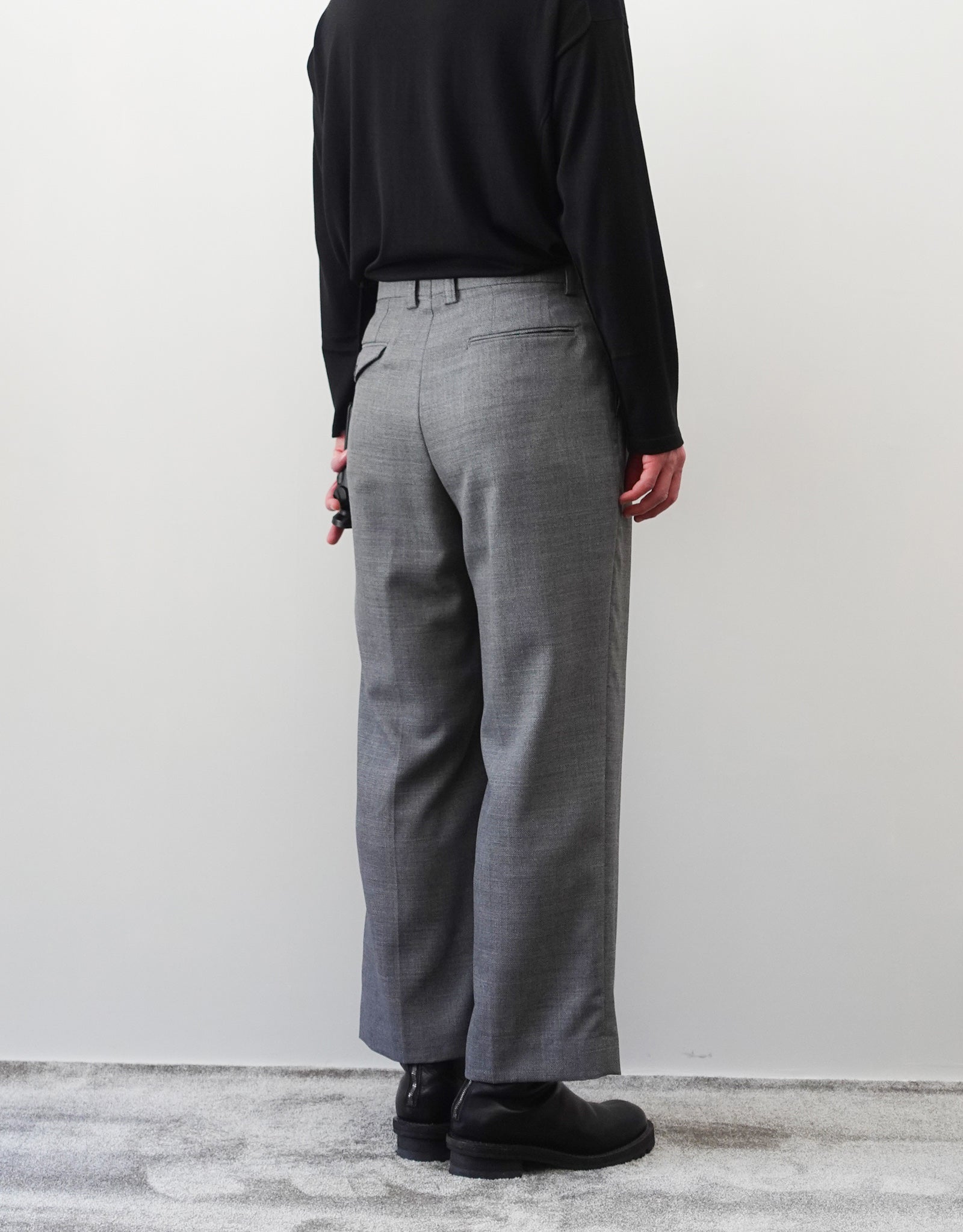 VOUILLON（ヴイヨン）のFour Pleats Wide Trousers - Gray（4プリーツワイドトラウザーズ）。ソリッドなグレーの生地に、腰元の4プリーツが深みのある陰影を生み出すワイドパンツ。洗練されたミニマルなスタイリング。モデル着用左後ろ向き画像。