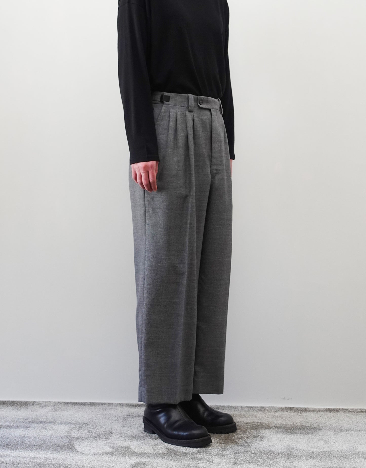 VOUILLON（ヴイヨン）のFour Pleats Wide Trousers - Gray（4プリーツワイドトラウザーズ）。ソリッドなグレーの生地に、腰元の4プリーツが深みのある陰影を生み出すワイドパンツ。洗練されたミニマルなスタイリング。モデル着用左向き画像。