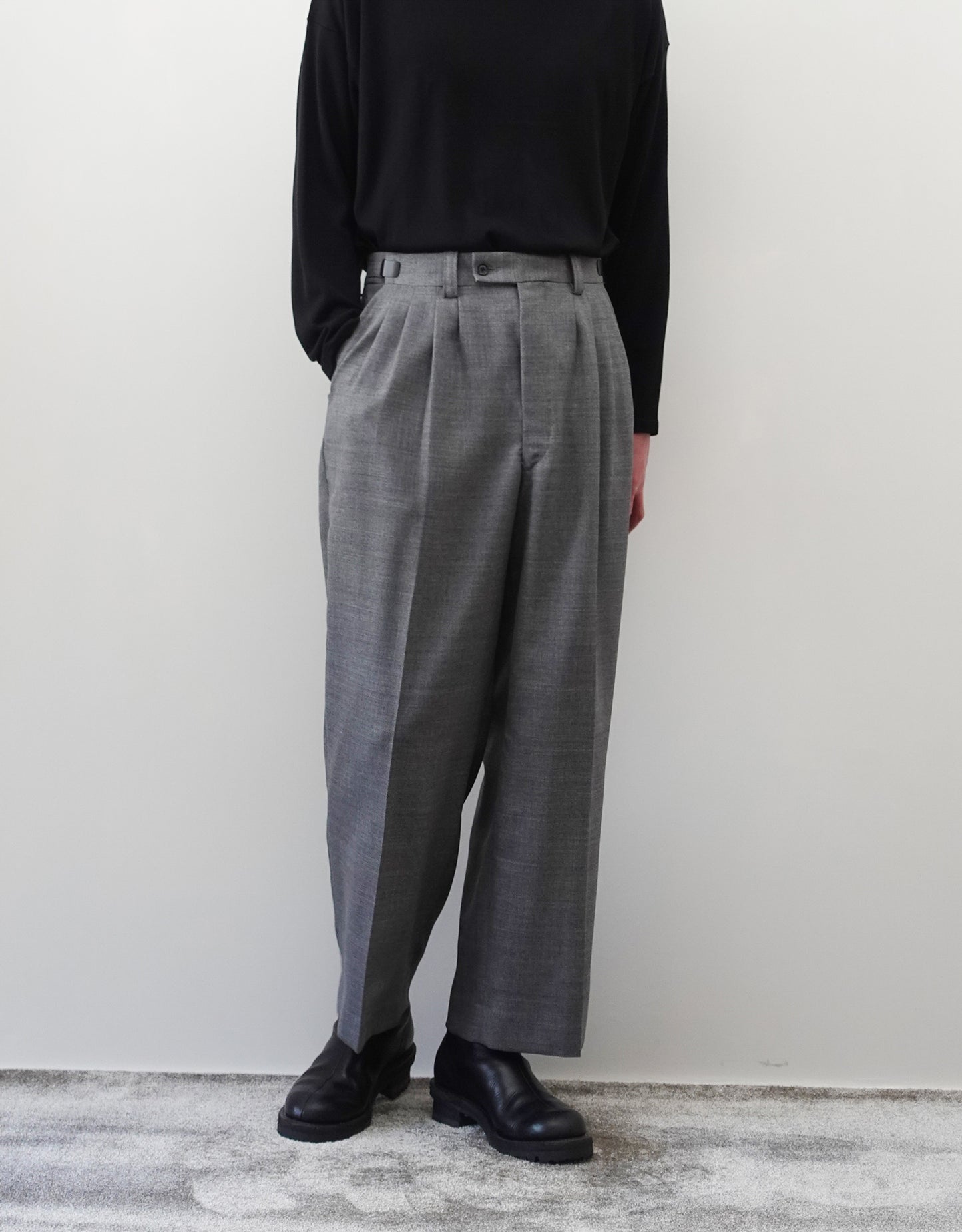 VOUILLON（ヴイヨン）のFour Pleats Wide Trousers - Gray（4プリーツワイドトラウザーズ）。ソリッドなグレーの生地に、腰元の4プリーツが深みのある陰影を生み出すワイドパンツ。洗練されたミニマルなスタイリング。モデル着用正面右ポケットに手を入れている画像。