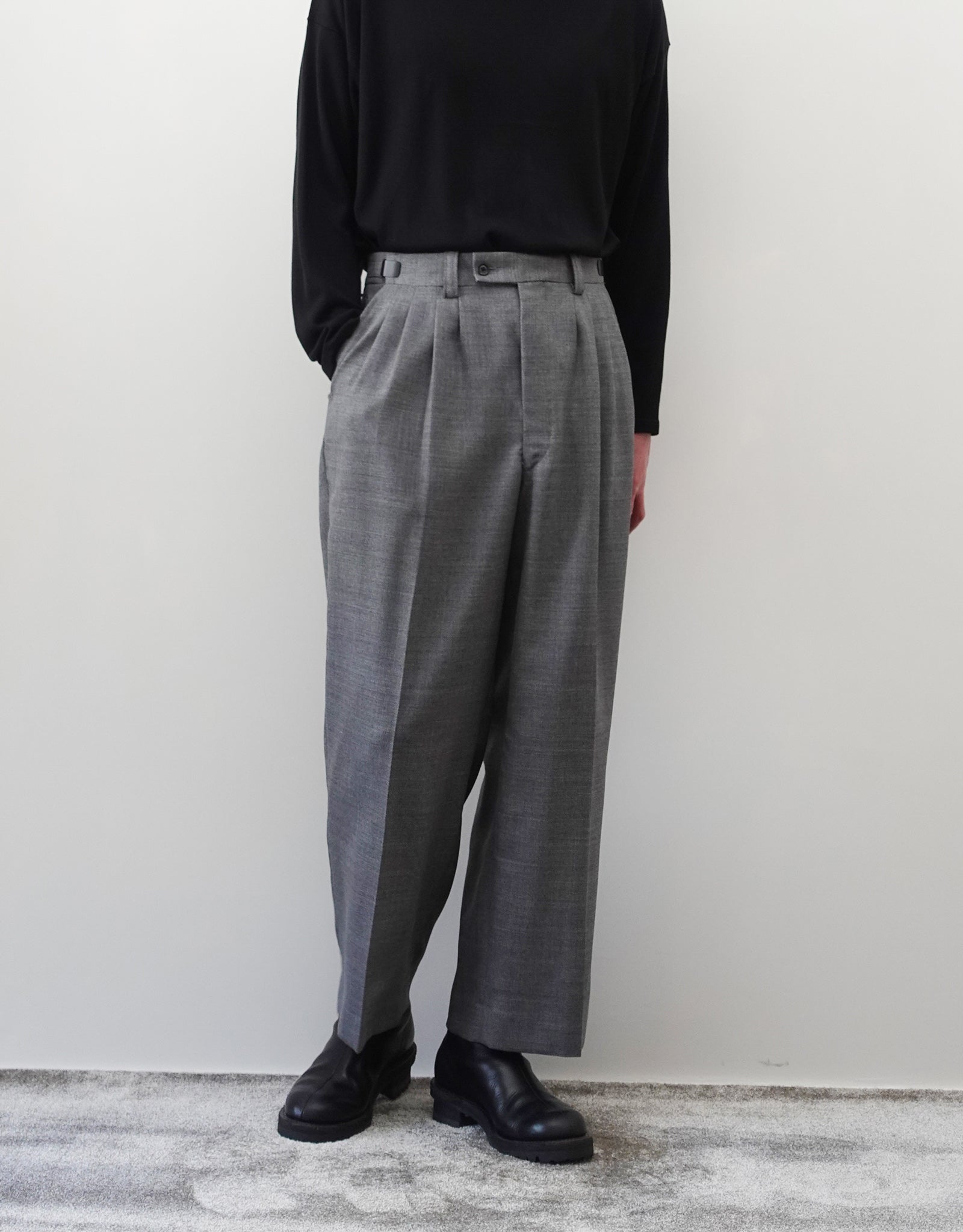 VOUILLON（ヴイヨン）のFour Pleats Wide Trousers - Gray（4プリーツワイドトラウザーズ）。ソリッドなグレーの生地に、腰元の4プリーツが深みのある陰影を生み出すワイドパンツ。洗練されたミニマルなスタイリング。モデル着用正面右ポケットに手を入れている画像。