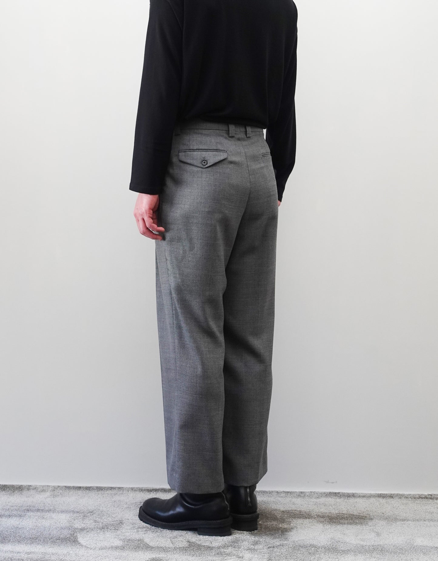 VOUILLON（ヴイヨン）のFour Pleats Wide Trousers - Gray（4プリーツワイドトラウザーズ）。ソリッドなグレーの生地に、腰元の4プリーツが深みのある陰影を生み出すワイドパンツ。洗練されたミニマルなスタイリング。モデル着用右後ろ向き画像。