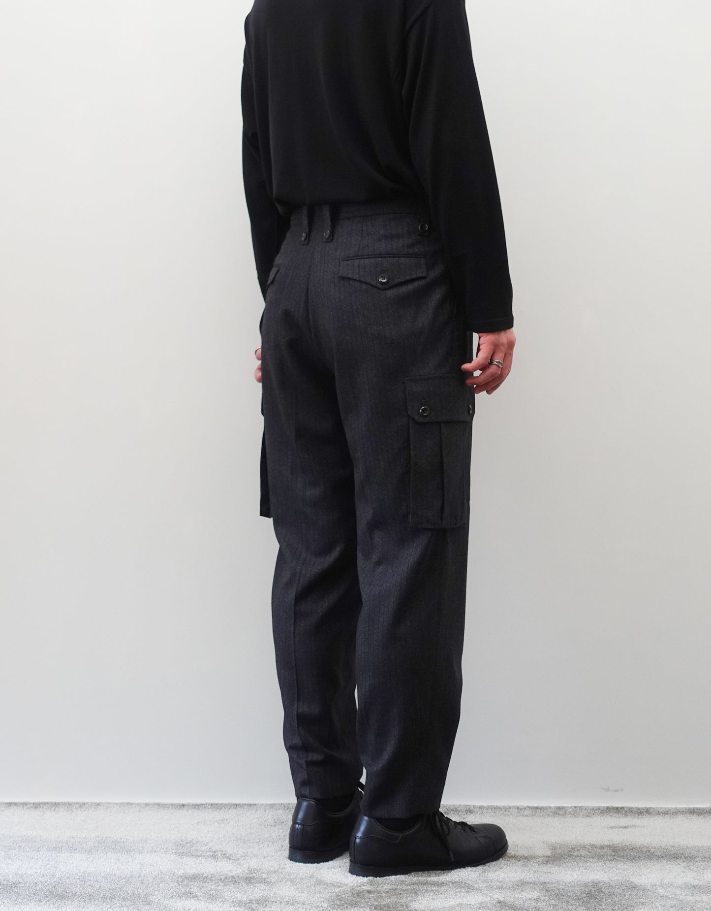 VOUILLON（ヴイヨン）のTwo Tuck Cargo Slacks - chacoal Stripe（2タックカーゴスラックス - チャコールストライプ）。上質な生地で立体的に仕上げることでシルエットの美しさとカーゴポケットの機能性の両立させたカーゴパンツ。モデル着用左斜め後向き画像。